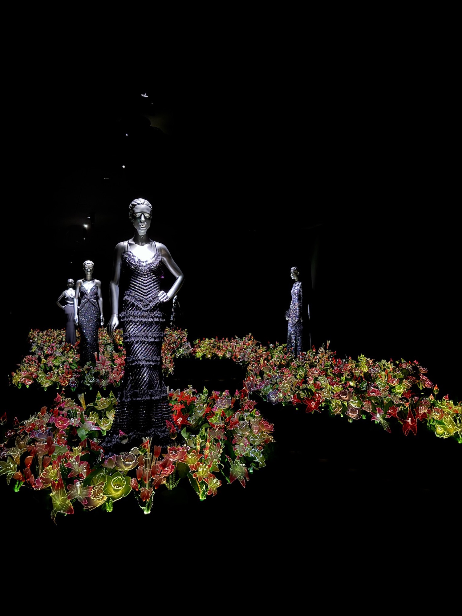 A museum fashion exhibit with mannequins in gowns surrounded by illuminated flowers